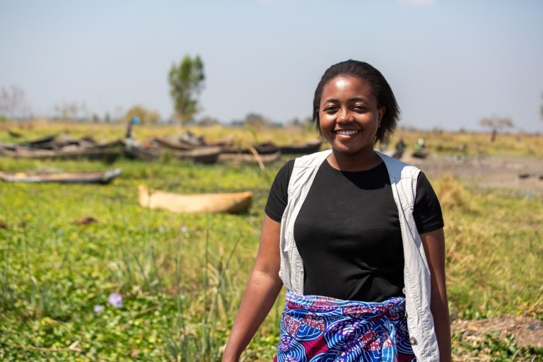 A young woman wearing a black tshirt and light colored vest stands outside with Lake Malawi in the background. Credit: Brian Clark/Pact.