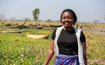 A young woman wearing a black tshirt and light colored vest stands outside with Lake Malawi in the background. Credit: Brian Clark/Pact.
