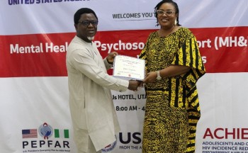 A man and woman stand in front of a red and white banner both holding a certificate between them and smiling at the camera.