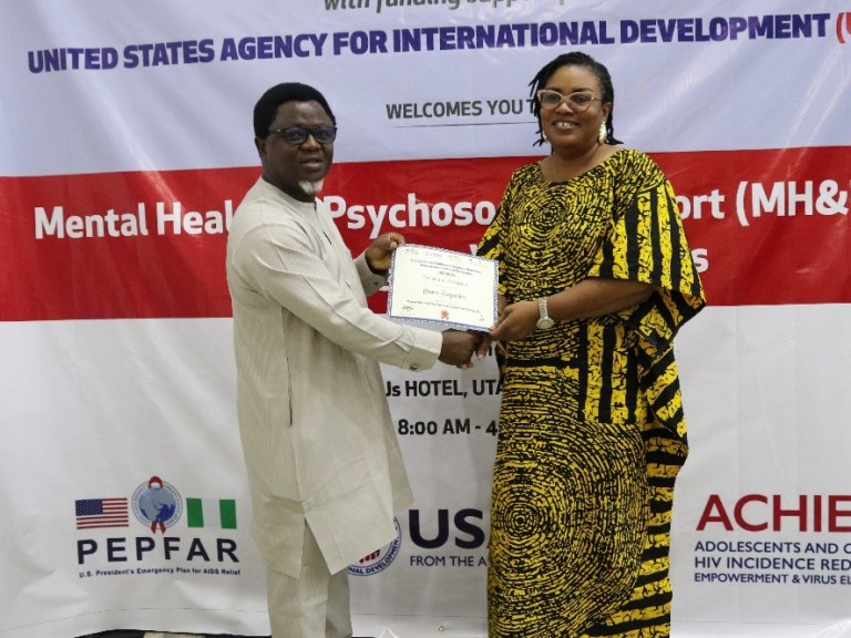 A man and woman stand in front of a red and white banner both holding a certificate between them and smiling at the camera.