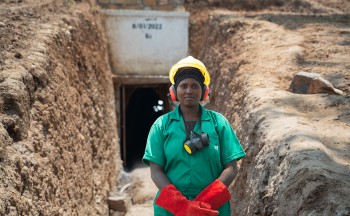 Christine Uwamahoro, a miner at GMDC who is also in charge of safety in the tunnels, stands outside in front of the entrance to a mining tunnel. She is wearing red gloves, a yellow hard hat, and a turquoise short sleeved uniform. Credit: Brian Clark/Pact.
