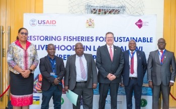 Six honored guests pose for a photo in front of a banner for the Restoring Fisheries for Sustainable Livelihoods in Lake Malawi project.