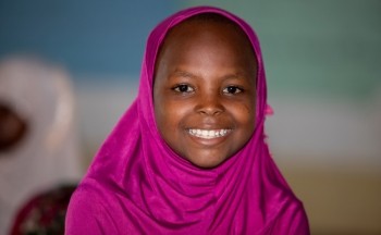 A young girl wearing a pink headscarf looks at the camera and smiles.