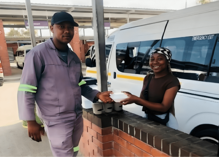 A young woman poses outside a taxi stand handing goods from her coffee business to a male customer.
