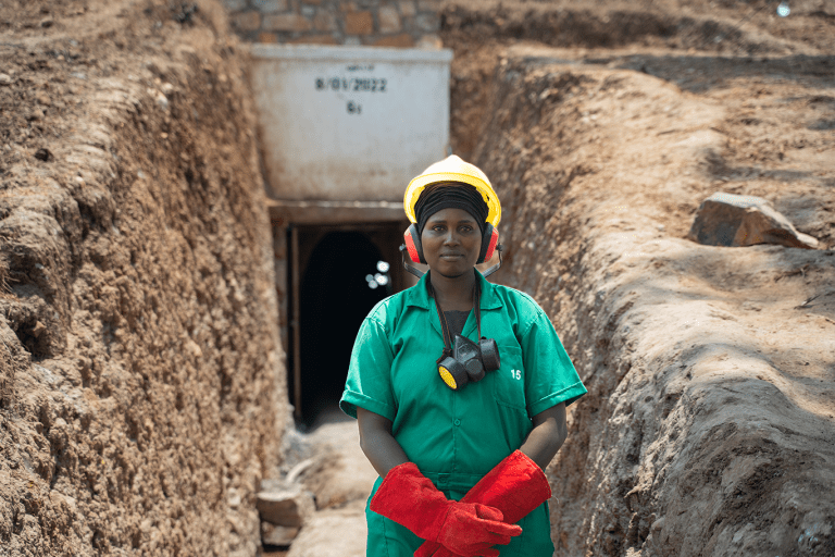 Christine Uwamahoro, a miner at GMDC who is also in charge of safety in the tunnels, stands outside in front of the entrance to a mining tunnel. She is wearing red gloves, a yellow hard hat, and a turquoise short sleeved uniform. Credit: Brian Clark/Pact.